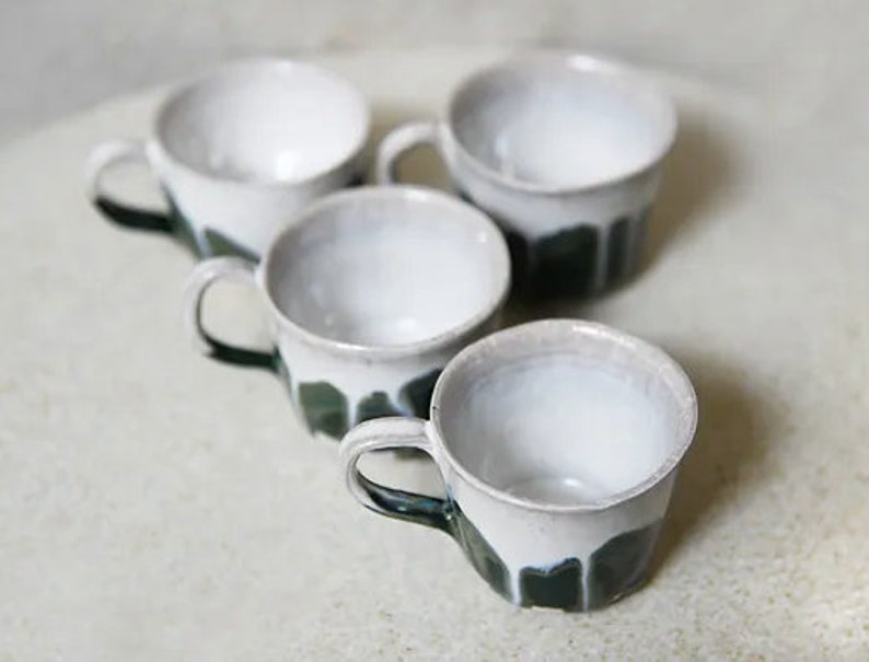 May include: A set of four ceramic mugs featuring a white glazed interior and upper exterior, transitioning into a dark green drip glaze pattern on the lower half. Each mug has a single curved handle and a slightly irregular, organic rim shape.
