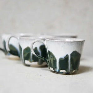 May include: A set of ceramic mugs featuring a white upper section and a dark green lower section with a distinctive drip glaze pattern. Each mug has a curved handle and a wide rim, showcasing a handcrafted aesthetic with visible colour blending.