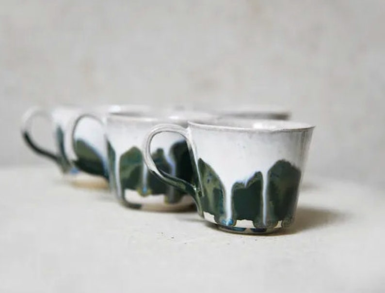 May include: A set of ceramic mugs featuring a white upper section and a dark green lower section with a distinctive drip glaze pattern. Each mug has a curved handle and a wide rim, showcasing a handcrafted aesthetic with visible colour blending.