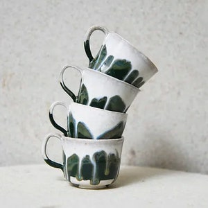 May include: A stack of four ceramic mugs with a white glaze on the upper portion and a dark green drip glaze pattern extending towards the base. Each mug features a curved handle on the side, and they are balanced in a vertical, slightly tilted column.