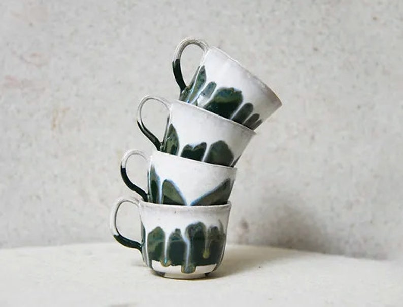 May include: A stack of four ceramic mugs with a white glaze on the upper portion and a dark green drip glaze pattern extending towards the base. Each mug features a curved handle on the side, and they are balanced in a vertical, slightly tilted column.