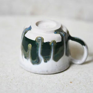 May include: A ceramic mug turned upside down, featuring a white base glaze with a dark green drip glaze pattern cascading from the top rim. The mug has a curved handle on the right side and a circular foot ring visible on the base.