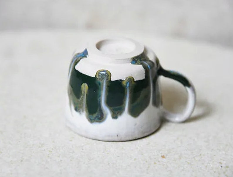 May include: A ceramic mug turned upside down, featuring a white base glaze with a dark green drip glaze pattern cascading from the top rim. The mug has a curved handle on the right side and a circular foot ring visible on the base.