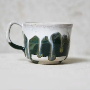 May include: A ceramic mug featuring a white upper section and a dark green lower section. The glaze is applied in a drip pattern, where the white glaze runs down over the dark green base, creating vertical streaks and a textured, layered appearance.