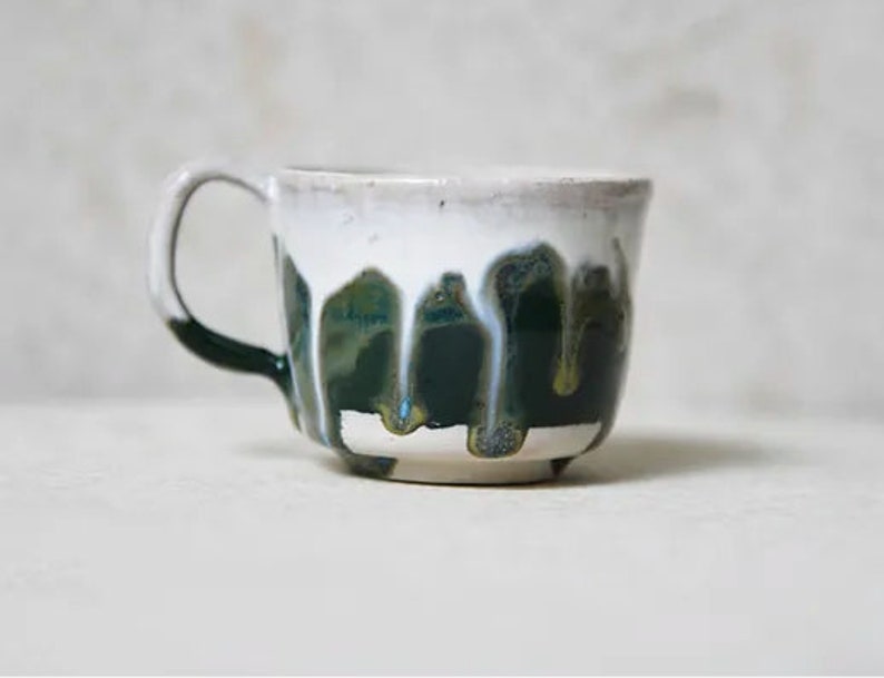 May include: A ceramic mug featuring a white upper section and a dark green lower section. The glaze is applied in a drip pattern, where the white glaze runs down over the dark green base, creating vertical streaks and a textured, layered appearance.