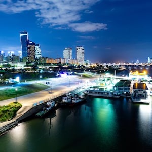 May include: Nighttime cityscape with illuminated skyscrapers and buildings along a waterfront. Several boats and ferries are docked, reflecting lights on the water. The sky is a deep blue with scattered clouds.