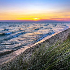 May include: A scenic beach at sunset. The ocean waves roll towards the sandy shore, with tall green grass on the right. The sky transitions from yellow to orange and purple.