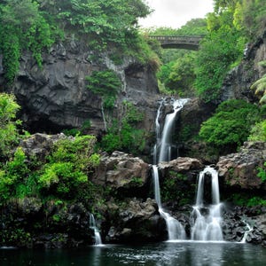 May include: A multi-tiered waterfall cascades over dark, jagged volcanic rock formations surrounded by lush green tropical foliage and dense forest. A stone bridge spans the top of the gorge above the flowing water, which pools at the base.
