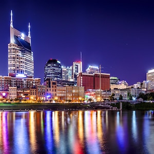 May include: Nighttime cityscape of Nashville, Tennessee, with illuminated skyscrapers and buildings reflecting in the water. The sky is a deep blue, and the water reflects the city lights in vibrant colors.