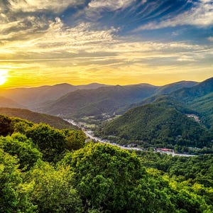 May include: A scenic landscape photograph of a mountain range at sunset. The sky is a gradient of yellow, orange, and blue, with the sun setting behind the mountains. Lush green trees and foliage are in the foreground.
