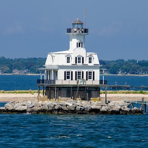 May include: A white lighthouse with black shutters and a lantern room sits on a rocky foundation surrounded by blue water. The structure features a multi-level design with a balcony and a small pier extending from the base into the calm sea.