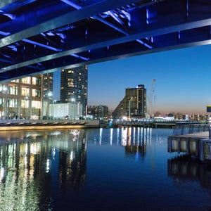 May include: A nighttime cityscape featuring a bridge with blue lighting, reflecting in the dark water below. Buildings with illuminated windows line the waterfront, and the sky transitions from blue to a darker hue.