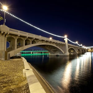 May include: Nighttime view of a concrete bridge with arched supports, illuminated by streetlights and a string of lights. The bridge spans a dark body of water, reflecting the lights. A second bridge is visible in the distance, with green lights.