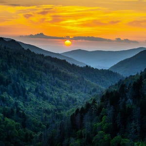 May include: A scenic landscape of mountain ranges covered in lush green forests under a vibrant sunset. The sky transitions from golden yellow to orange, with the sun peeking through the clouds.