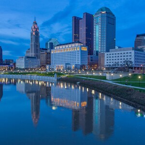 May include: The Columbus, Ohio skyline at twilight features a mix of modern and historic skyscrapers reflecting in the Scioto River. A green park with illuminated pathways runs along the riverbank, contrasting with the blue evening sky.
