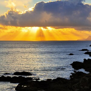 May include: A scenic coastal landscape at sunset. The sky is filled with golden light breaking through dark clouds, casting rays over the calm ocean. Rocky shoreline in the foreground, with mountains visible in the distance.