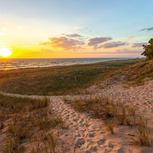 May include: A scenic landscape featuring a sandy beach and dunes, with a path leading towards the ocean. The sky is a gradient of yellow and blue, with the sun setting over the water. Green vegetation dots the dunes.