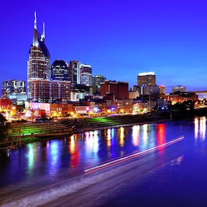 May include: A nighttime cityscape of Nashville, Tennessee, with illuminated skyscrapers and buildings reflecting in the dark blue water. A boat leaves a white wake in the foreground, with streaks of light.
