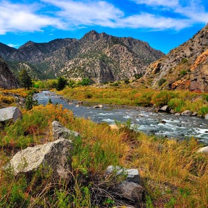 May include: A scenic landscape featuring a river winding through a canyon. The image showcases rocky mountains, lush greenery, and a bright blue sky with scattered clouds. The river's water is clear and flowing.