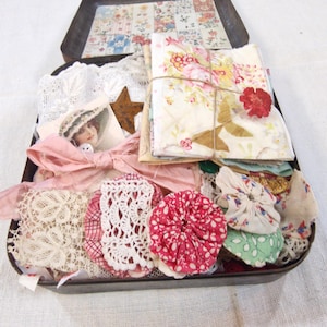 May include: A dark metal tin filled with assorted craft supplies including folded floral fabric bundles tied with twine, white lace pieces, fabric yo-yos in red and white patterns, a small vintage-style portrait card, and a rusty metal star.
