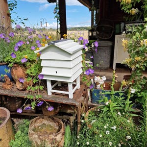 May include: A white wooden beehive with a sloped roof sits on a metal surface, surrounded by purple and yellow flowers. The hive has a small entrance hole and is supported by four white legs. The background includes a rustic porch and a field.