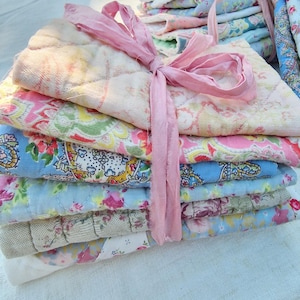 May include: A stack of folded, quilted fabric pieces featuring various floral and paisley patterns in shades of pink, blue, yellow, and white. The stack is secured with a light pink fabric ribbon tied in a bow around the centre of the textiles.