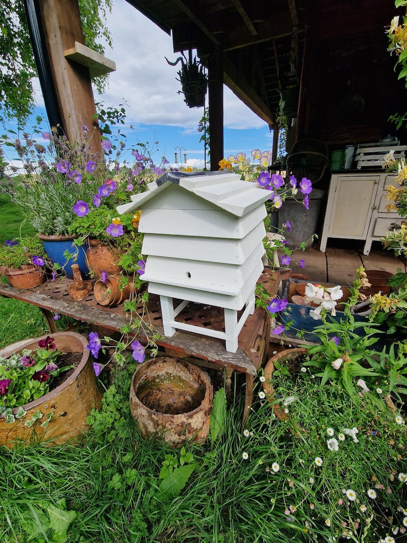 May include: A white wooden beehive with a sloped roof sits on a weathered metal bench, surrounded by potted plants and vibrant purple flowers. The hive has multiple tiers and a small entrance hole. The scene is set outdoors with a blue sky.