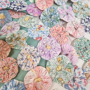 May include: A collection of numerous fabric yo-yos in various floral, plaid, and polka dot patterns, each featuring a gathered circular design with a small yellow centre. The fabric circles are arranged in a dense cluster on a light green base.