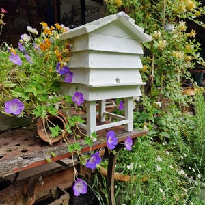 May include: A white wooden beehive with a gabled roof, set on a table with purple flowers and greenery. The hive has multiple tiers and a small entrance hole. The scene is outdoors, surrounded by plants and flowers.