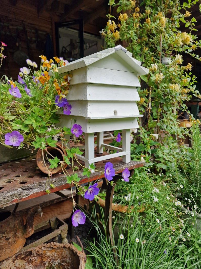 May include: A white wooden beehive with a gabled roof, set on a table with purple flowers and greenery. The hive has multiple tiers and a small entrance hole. The scene is outdoors, surrounded by plants and flowers.