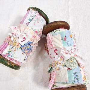 May include: Two vintage wooden spools wrapped in assorted fabric scraps featuring floral, plaid, and solid pastel patterns. Each spool is tied with a white fabric ribbon and decorated with small, circular fabric yo-yo embellishments.