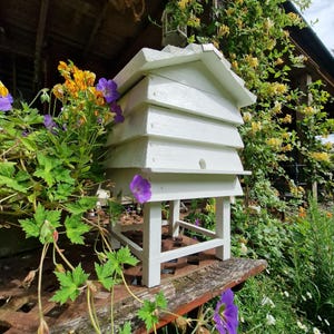 May include: A white wooden beehive with a tiered design, set on a stand. The hive is surrounded by vibrant purple and yellow flowers and lush green foliage. The scene is set outdoors, with a rustic wooden structure in the background.