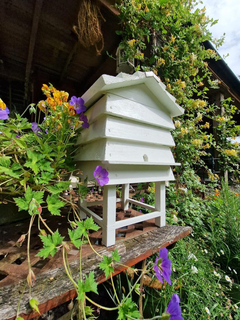 May include: A white wooden beehive with a gabled roof, set on a stand. The hive is surrounded by vibrant purple and yellow flowers and lush green foliage. The scene is set outdoors, with a rustic wooden structure in the background.