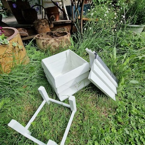 May include: A white wooden planter box with a matching stand and lid, resting on green grass. The box is rectangular and appears to be made of painted wood. The lid is partially open, and the stand is separate.