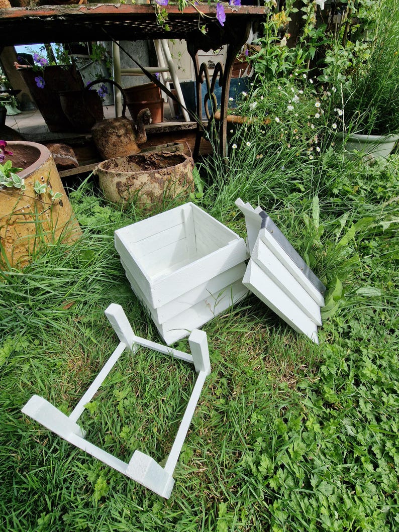 May include: A white wooden planter box with a matching stand and lid, resting on green grass. The box is rectangular and appears to be made of painted wood. The lid is partially open, and the stand is separate.