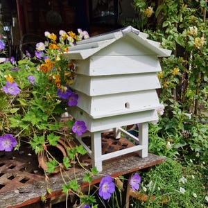 May include: A white wooden beehive with a tiered design, set on a small stand. The beehive is surrounded by vibrant purple and yellow flowers and lush green foliage, creating a natural garden setting.