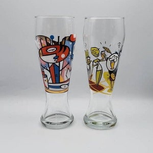 May include: Two clear pilsner glasses with colorful cartoon illustrations. One glass features a red and white character in a suit, the other depicts a group of people enjoying beer. The glasses have a flared shape and a sturdy base.