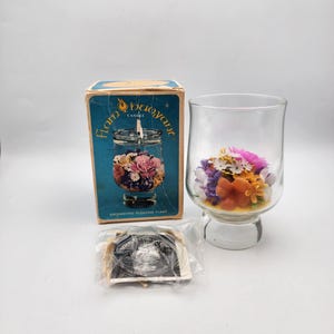 May include: A vintage "Flamboyant Candle" box with a glass candle holder filled with colorful wax flowers. The box has a blue background with white text and a picture of a similar candle holder. The candle holder is on a white surface.