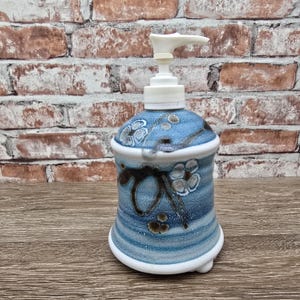 May include: A ceramic soap dispenser with a blue and white floral design. The dispenser has a white pump and a rounded shape with a flared base. The body of the dispenser features a hand-painted floral pattern and horizontal blue stripes.