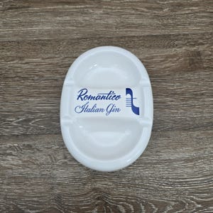 May include: White oval ashtray with two recessed areas and a central divider. The ashtray features the words "Romantico Italian Gin" in blue script. A blue graphic is on the right side of the text.