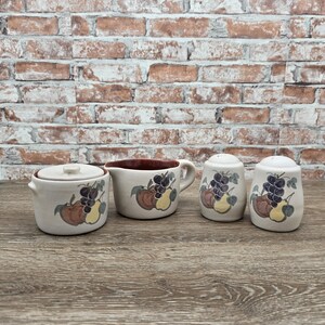 May include: A cream-colored ceramic kitchen set featuring a sugar bowl with a lid, a creamer, and two salt and pepper shakers. Each piece is decorated with a fruit design of grapes, apples, and pears in shades of purple, brown, and yellow.