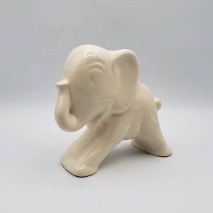 May include: A cream-colored ceramic elephant figurine. The elephant is depicted in a walking pose with its trunk raised. The figurine has a smooth, glossy finish and is set against a plain white background.