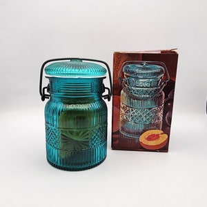 May include: A teal glass jar with a lid and a black metal handle. The jar has a ribbed and patterned design. A box with a similar jar illustration is next to it.