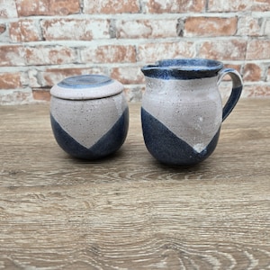 May include: A ceramic sugar bowl with a lid and a small pitcher. Both pieces are speckled with a light gray and navy blue geometric design. The pitcher has a handle and a slightly flared rim. The pieces are on a wooden surface.