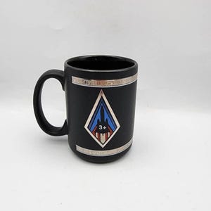 May include: A matte black ceramic mug featuring a silver-toned horizontal band at the top and bottom. The center displays a diamond-shaped emblem with a blue and red graphic of an SR-71 Blackbird aircraft and the text SR-71 BLACKBIRD.