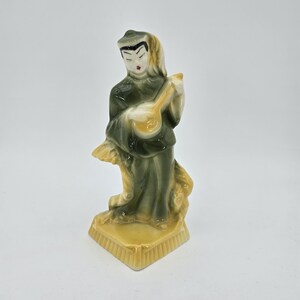 May include: A glazed ceramic figurine depicting a person in dark green robes playing a light yellow stringed instrument. The figure stands on a yellow base with a textured edge, featuring painted facial details and a head covering in a glossy finish.