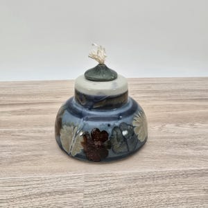 May include: A blue and white ceramic oil lamp with a floral design. The lamp has a round base and a small, green lid with a wick.