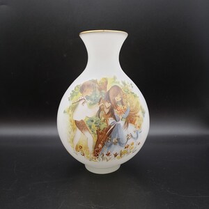 May include: Frosted glass vase with a gold rim, featuring a painted scene of two children in a garden. The artwork depicts a boy offering a heart to a girl holding flowers, with a backdrop of trees and flowers.