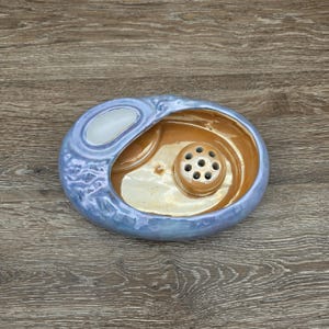 May include: An oval ceramic dish with a glossy iridescent light blue exterior and a smooth metallic gold interior. The dish features a raised circular center piece with seven small holes and a separate white oval indentation on the upper rim.