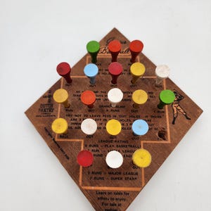 May include: A diamond-shaped wooden baseball peg game board featuring printed instructions, the text Dutch Pantry Family Restaurants, and various colored pegs including red, green, blue, yellow, and white inserted into the board holes.